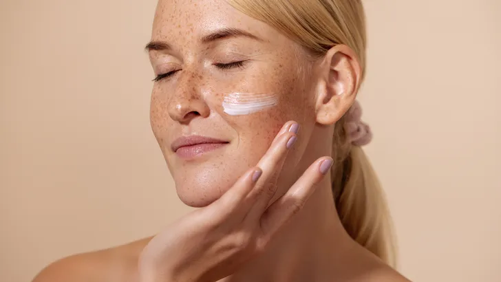 Close up of beautiful young female with cream on a face. Smiling woman with closed eyes touching cheek with fingers.