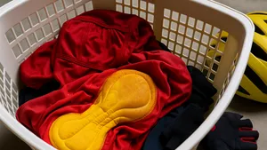 Natte rode wielerkoersbroek met felgele zeem en bijpassende jersey liggen verkreukeld in een witte wasmand, met handschoen en helm ernaast.