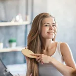 Portrait of joyful blonde lady brushing her long hair near looking glass at bathroom