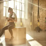 Staying in touch. Attractive athletic woman in headphones wearing towel on her shoulders and using smart phone while sitting at gym