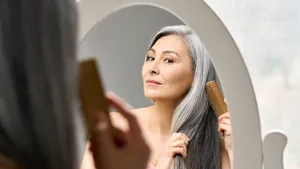 Een vrouw van midden 50 met grijs haar kijkt in de spiegel en kamt haar volle grijze haar.