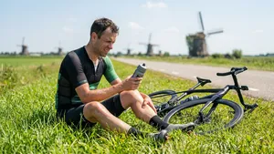 Gefrustreerde wielrenner zonder helm zit in het gras langs een polderweg en masseert zijn verkrampte kuit, met zijn racefiets naast zich.