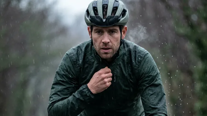 Wielrenner op de racefiets met zwarte beenstukken ritst zijn regenjack voor wielrennen open voor ventilatie op een nat wegdek.