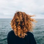 View from the back a woman with curly hair looks at the sea