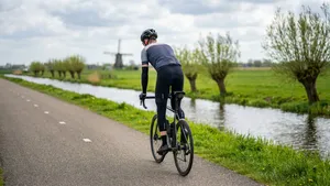 Mannelijke wielrenner met helm, armstukken en beenstukken staat op de pedalen in een Nederlands polderlandschap om zadelpijn te verlichten.