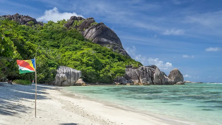 Tropische eilanden van Afrika – Foto van wit zandstrand met palmbomen en blauwe zee. Op het strand staat de vlag van de Seychellen.