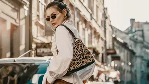 Outdoor autumn fashion portrait of elegant, luxury woman wearing sunglasses, trendy white shirt, leather trousers, with animal, leopard print bag, walking in street of European city. Copy, empty space