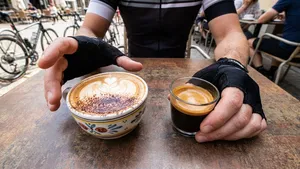 Een close-up van de handen van een wielrenner met fietshandschoenen aan, die op een cafétafel twijfelt tussen een grote, romige cappuccino aan de ene kant en een kleine, krachtige espresso aan de andere kant.