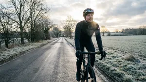 Een wielrenner in volledige winterkleding lacht terwijl hij op een racefiets rijdt over een koude, natte weg langs bevroren velden. Hij kijkt omhoog en geniet van de omgeving.