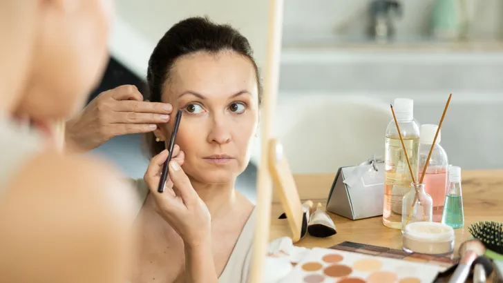 Een rustige vrouw van middelbare leeftijd brengt haar oogmake-up aan terwijl ze in de spiegel kijkt, zittend aan een tafel in haar huis.