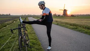 Wielrenner met helm, armstukken en beenstukken rekt zijn hamstrings door zijn been op een hek te leggen, met zijn racefiets en een molen in een polderlandschap bij zonsondergang op de achtergrond.