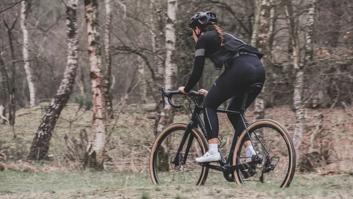 Vrouwelijke wielrenner met helm rijdt op een aero gravelbike met extreem brede mountainbikebanden over een Nederlands gravelpad langs een dijk. Op de achtergrond weilanden, een kanaal en een windmolen.