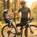 Vader in fietskleding geeft lachend een high-five aan zijn kind in een kinderzitje op de bagagedrager van een gravelbike in een zonnig bos.
