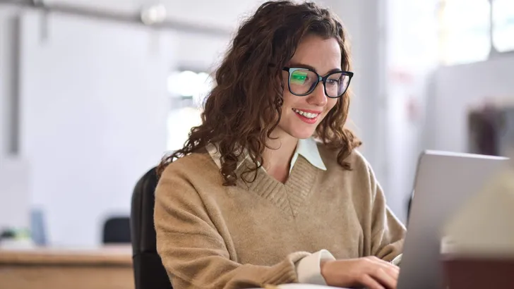 Young happy woman employee or student sitting at desk using laptop.