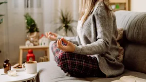 Cozy woman in knitted winter warm sweater and checkered pajama drinking hot cocoa or coffee in mug, reading book, meditation yoga on couch at home. Christmas holidays with decor and lights.