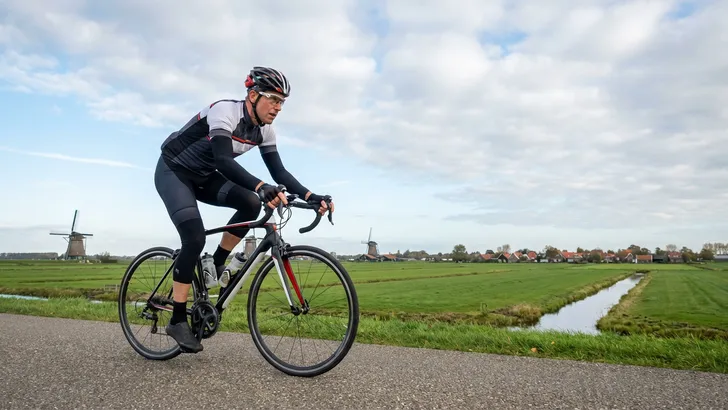 Een mannelijke wielrenner op een racefiets in een dynamische en efficiënte fietshouding op een landweg, die het resultaat van een correcte zadelhoogte illustreert.