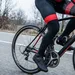Close-up van een mannelijke wielrenner in winterkleding, inclusief zwarte lange fietsbroek met rode accenten en overschoenen, die op een racefiets rijdt op een koude, bewolkte dag langs een weg met kale bomen. Zijn ademwolk is zichtbaar.