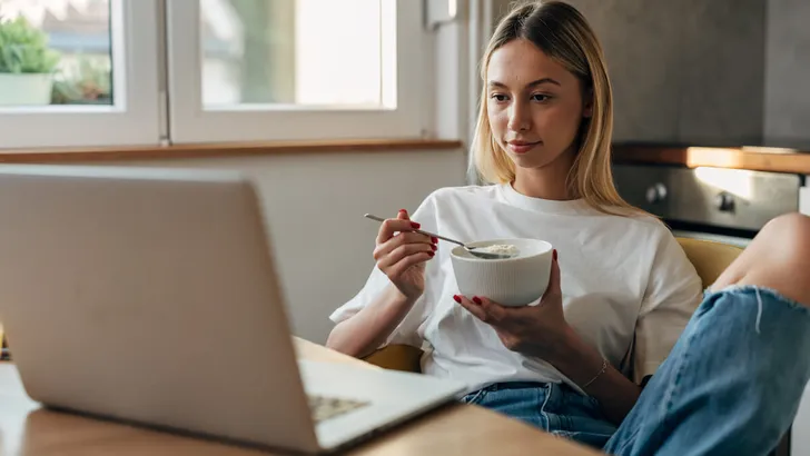 Young woman eating oatmeal in the kitchen and looking at the laptop.
