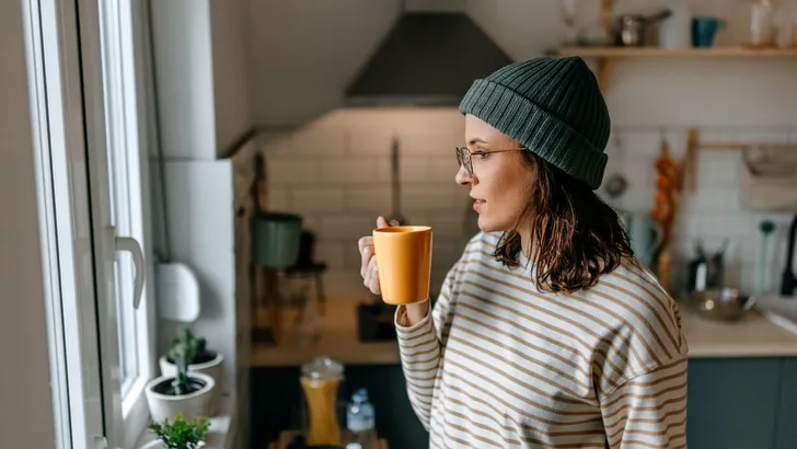 Woman with coffee cup looking through window at home