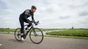 Een mannelijke wielrenner in volledige uitrusting fietst staand op de pedalen op een vlakke weg in de Nederlandse polder.