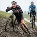 Amateurvelderijder glijdt met een gezicht vol angst en pijn een steile, modderige dijk af tijdens een cyclocross wedstrijd. Achter hem rijdt een professional in een Belgische trui soepel door.