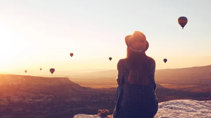 View from the back of a girl in a hat sits on a hill and looks at air balloons.