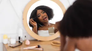 Young Afro lady applying mascara on lashes near mirror, speaking on smartphone, hurrying to work, being late at home