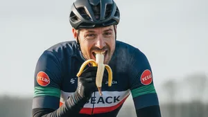 Een krachtige wielrenner met een winterjas en helm fietst door een Nederlands winterlandschap terwijl hij een banaan eet als sportvoeding.
