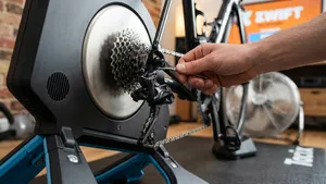 Gedetailleerde close-up van een achterderailleur en cassette van een racefiets op een fietstrainer, waarbij een hand de stelschroef aanpast om de versnellingen af te stellen.