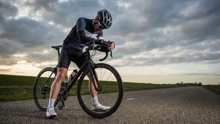 Een uitgeputte mannelijke wielrenner met helm en fietskleding hangt over het stuur van zijn racefiets, stilstaand op een polderweg onder een bewolkte lucht.