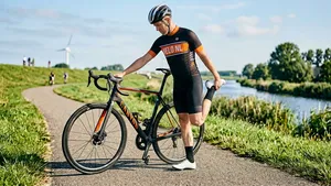 Een mannelijke wielrenner in fietskleding staat zonder helm naast zijn racefiets op een parkeerplaats en rekt zijn bovenbeenspier door zijn hak tegen zijn billen te trekken, als warming-up voor een fietstocht.