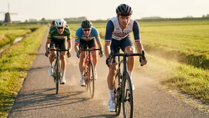 Een gefocuste wielrenner rijdt zijn zwoegende vrienden uit het wiel tijdens een intensieve rit op een zonnige polderweg met een molen in de achtergrond.