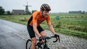 Een vermoeide amateurwielrenner in een oranje-zwart Wieler Revue shirt zwoegt op zijn racefiets over een nat kasseienpad in het Nederlandse polderlandschap, met een molen op de achtergrond.