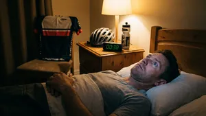 Een man ligt midden in de nacht klaarwakker en gefrustreerd in bed te staren naar het plafond. Op het nachtkastje naast hem staan een fietshelm en een bidon, en op de achtergrond hangt een wielershirt, wat duidt op een recente avondtraining.