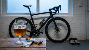 Een racefiets staat naast een glas speciaalbier op een houten tafel, wat de tegenstelling tussen sport en ontspanning weergeeft.