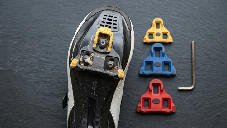 Een flat lay-afbeelding met een realistische witte wielerschoen, drie gekleurde schoenplaatjes (geel, blauw, rood), en een inbussleutel op een donkere achtergrond.