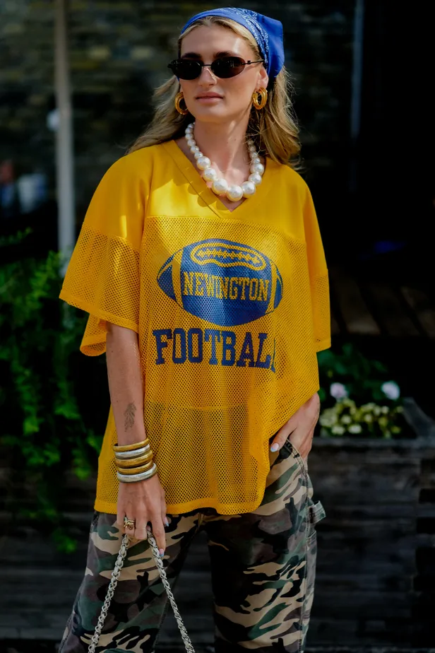 Vrouw met zonnebril en lang blond haar, geel football shirt en blauw sjaaltje in haar 