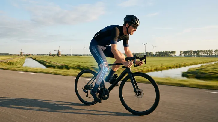 Mannelijke wielrenner met helm fietst over een polderweg, met een artistiek röntgeneffect dat zijn beenbotten subtiel toont.