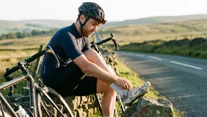 Mannelijke wielrenner met helm zit langs de weg en kijkt peinzend naar zijn versleten wielerschoen, een teken dat het tijd is om fietsschoenen te vervangen.