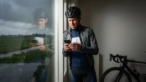 Wielrenner in fietskleding checkt gespannen Buienradar op smartphone bij een raam, met dreigende regenwolken buiten en zijn racefiets op de achtergrond.