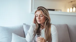 Een vrouw zit op de bank met een glas water, wachtend tot haar hoofdpijn afneemt.