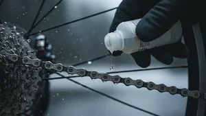 Close-up van een natte ketting en cassette van een racefiets terwijl met een flesje smeermiddel één druppel op de ketting wordt aangebracht, winterse omstandigheden.