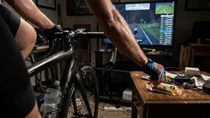 Vanuit het perspectief van de fietser: een bezwete hand grijpt naar een bidon op een indoor trainer in een sfeervol verlichte 'pain cave'.