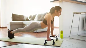 Young woman doing sport workout in room during quarantine. Picture of strong fitness model stand in plank position using push up stands hand bar. Exercising in room alone.