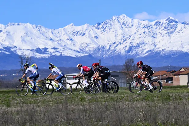 Een peloton wielrenners rijdt door een zonnig Italiaans landschap met de besneeuwde toppen van de Alpen op de achtergrond tijdens de koers Milaan-Turijn.  