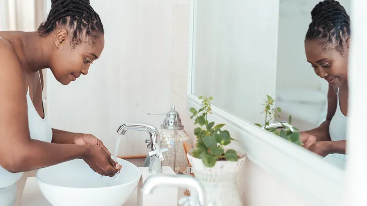 Young African woman standing over the sink in her bathroom washing her face with water in the morning