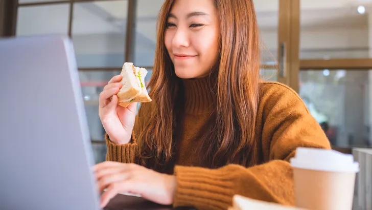 An asian woman holding and eating whole wheat sandwich while working on laptop computer