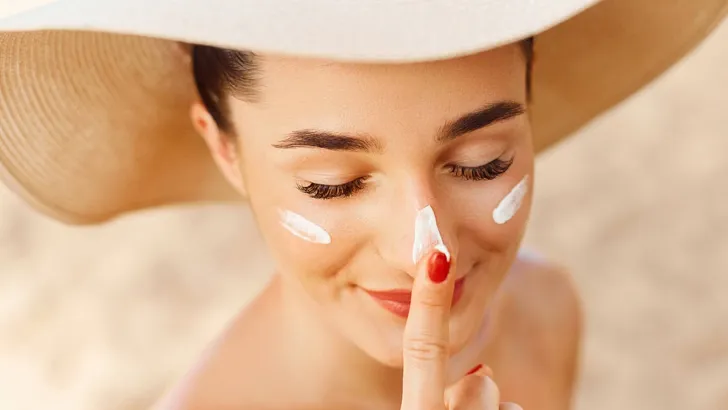 Beautiful Young woman with sun cream on face. Girl holding sunscreen bottle on the beach. Female in hat applying  moisturizing lotion on skin.Skin care. Sun protection. Suntan