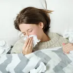 A young woman lies in bed under a blanket and blows her nose in