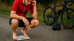 Wielrenner in fietskleding staat na de rit balend op een weegschaal, met zijn koersfiets op de achtergrond.